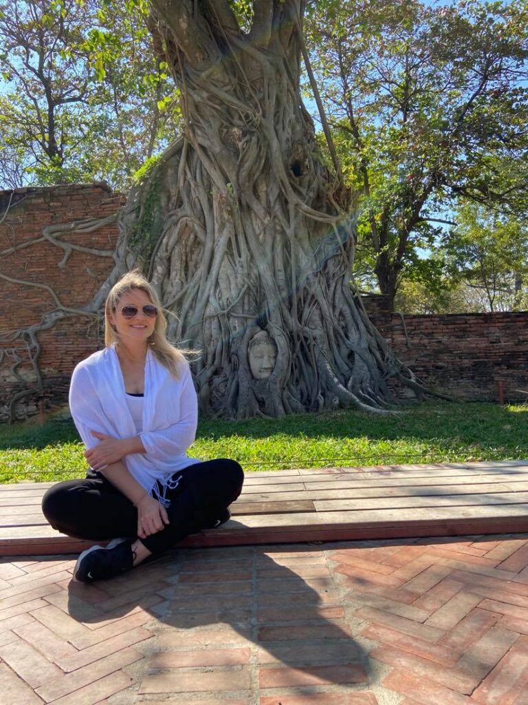 Image of the author, Lynn Howard, sitting under a banyan tree with a buddha's head in the roots.